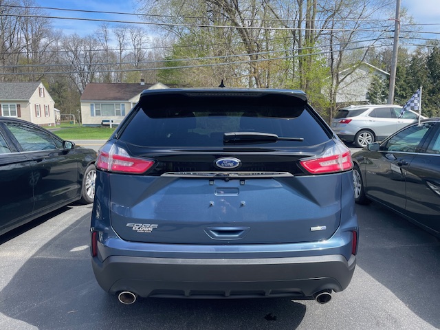 Rear view of a blue Ford Edge SUV in a parking lot, showing taillights, Ford emblem, Edge badge, rear wiper, and dual exhausts.