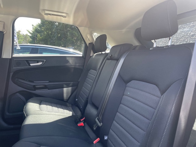 Back seat of a car with black fabric upholstery, three headrests, and visible seat belts by the doors and windows sunlit outside.