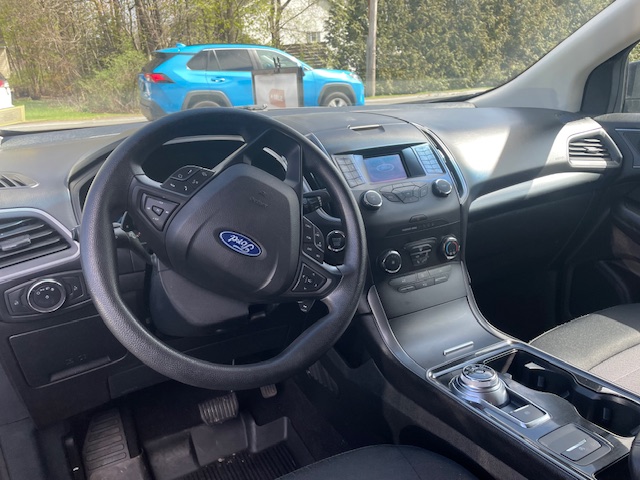 Interior of a Ford with steering wheel, center console featuring a rotary gear dial, touchscreen display, and dashboard controls; a blue SUV is outside through the windshield.