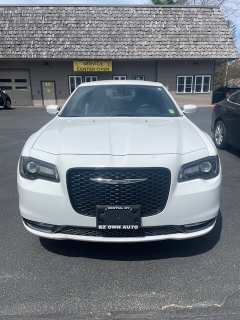 Front view of a white SUV with a black mesh grille and Chrysler emblem, parked in a dealership lot with a service center sign in the background.