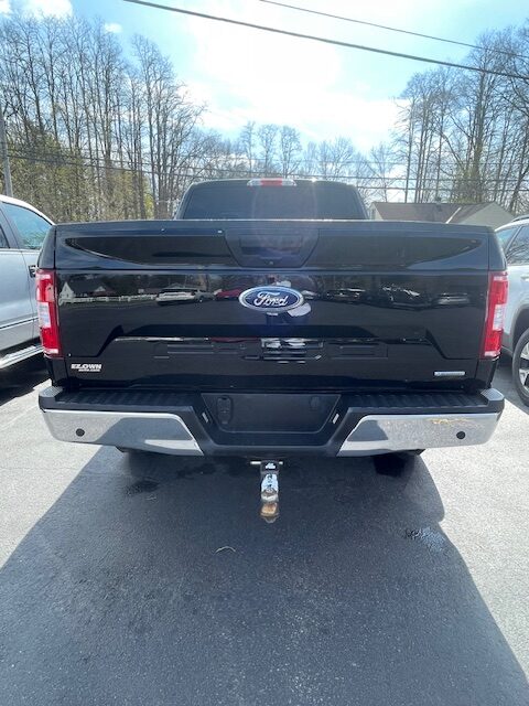 Rear view of a black Ford pickup truck with a trailer hitch, parked in a lot with other cars and trees in the background.