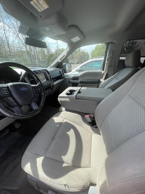 Front interior of a Ford pickup: gray fabric seats, steering wheel, and center console with cup holders visible in daylight.