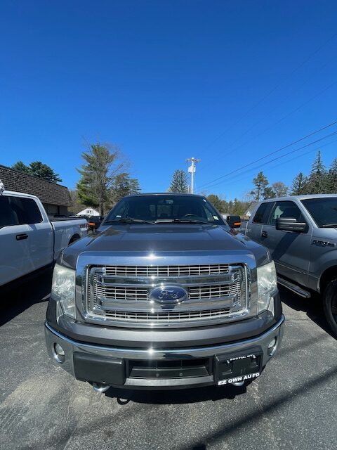 Front view of a blue Ford pickup truck in a dealership lot, flanked by white and silver trucks under a clear blue sky.