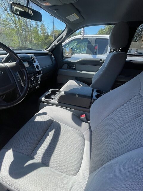 Front cabin of a gray pickup truck with cloth seats, center console and cupholders, steering wheel on the left; sunlight streaming in and another vehicle visible outside.