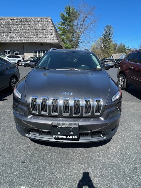 Front view of a dark gray Jeep Grand Cherokee SUV facing the camera in a dealership lot with a seven-slot grille and clear blue sky.