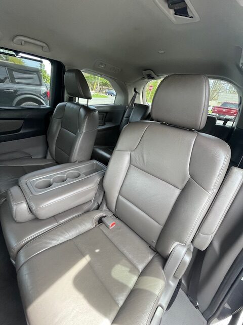 Interior of a vehicle featuring tan leather captain's chairs in the second row, with a folded-down center armrest and cup holders; front and back rows visible.