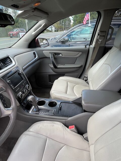 Beige leather SUV interior showing front seats, center console with gear shifter and cup holders, and dashboard screen; outside cars and an American flag visible through the window.
