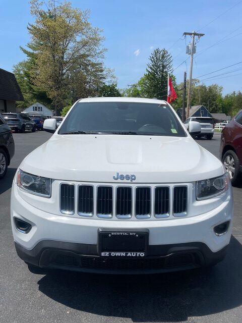 Front view of a white Jeep SUV parked in a dealership lot, featuring the signature seven-slot grille, rectangular headlights, and an EZ OWN AUTO license plate frame.