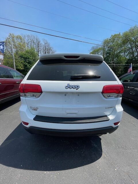 Rear view of a white Jeep SUV parked in a dealership lot, showing the Jeep badge, rear spoiler, and taillights with trees and other cars in the background.