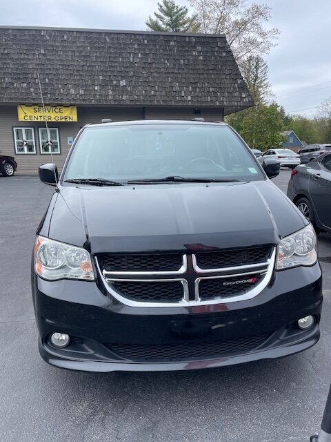 Front view of a dark Dodge minivan parked in a lot, headlights and chrome grille visible, service center building in the background.