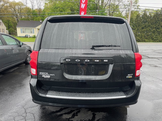 Back view of a black Dodge Grand Caravan in a wet parking lot, showing rear hatch, taillights, rear window wiper, and badges on the left reading 'Grand Caravan'.