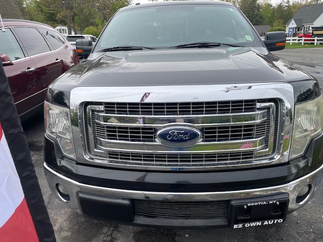 Front view of a gray Ford pickup truck with a large chrome grille and blue Ford emblem, parked in a lot beside a red SUV; dealership plate frame reads EZ OWN AUTO.