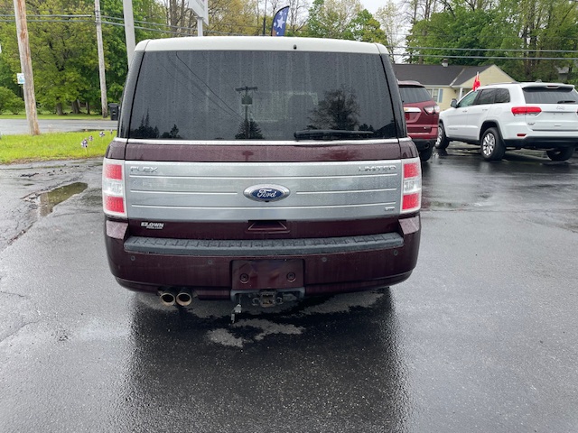Rear view of a maroon Ford Flex Limited parked in a wet parking lot; dual exhaust pipes visible and the license plate mount is empty, with other vehicles and trees in the background.
