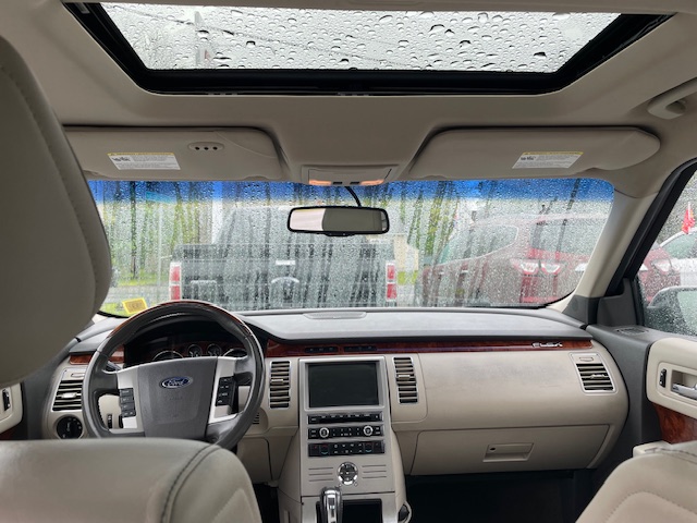 Inside a beige Ford SUV, view of the dashboard, center touchscreen, wood trim, and sunroof with raindrops on the windshield