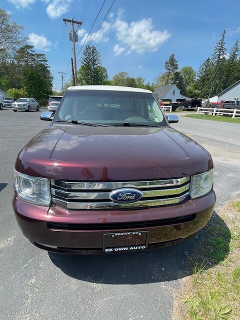 Front view of a maroon Ford SUV with a white roof parked on a paved lot, blue sky and trees in the background.