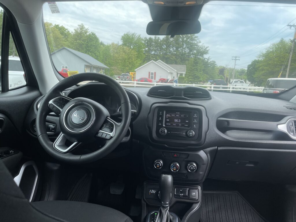 Driver's-seat view of a Jeep with steering wheel, center console, and gear shifter; outside shows trees, houses, and parked cars through the windshield.