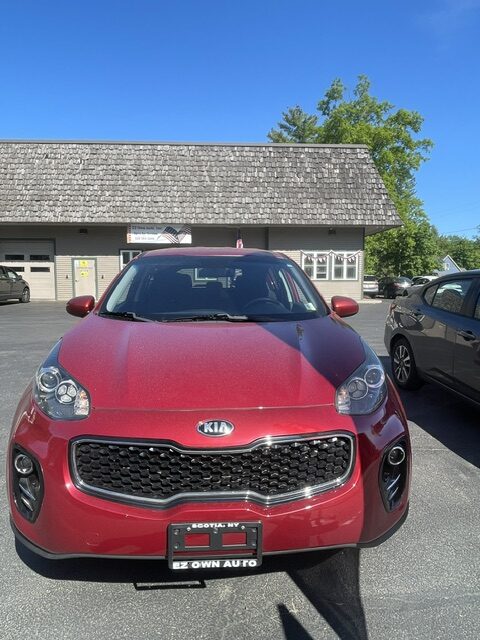 Front view of a red Kia SUV parked in a lot in front of a gray building on a sunny day with a clear blue sky.