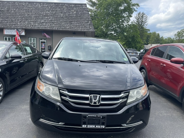Front view of a black Honda minivan in a dealership lot, with other cars and a small building in the background.