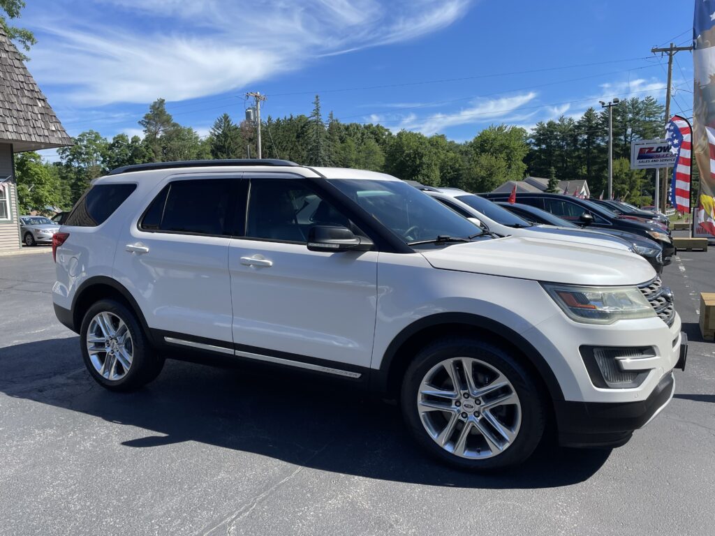 White SUV in a car dealership lot with other vehicles, blue sky and trees in the background, and a dealership sign visible on the right side
