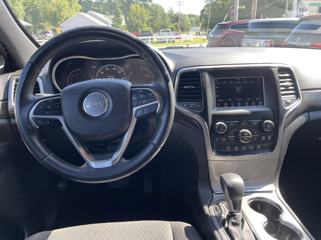 Jeep driver’s cockpit: steering wheel, instrument cluster, center touchscreen, and gear selector; parked outside in a lot.
